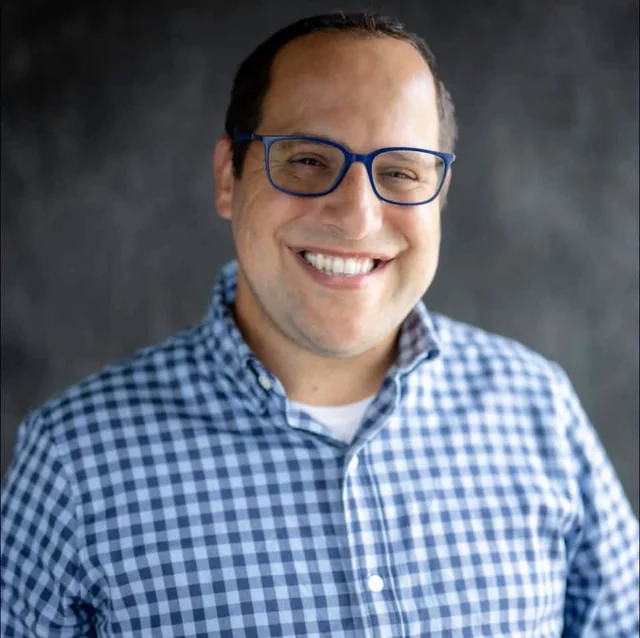 Eli Lebowicz smiling in blue glasses and a blue checked shirt